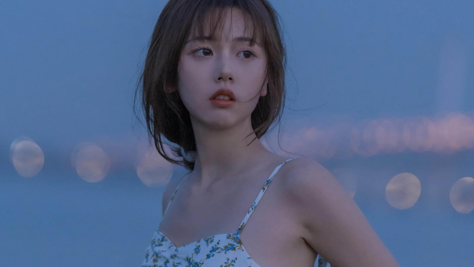 꽃무늬 캐미솔 원피스를 입은 성인 여성이 푸르게 흐릿한 배경 앞에서 옆을 바라보고 있다.