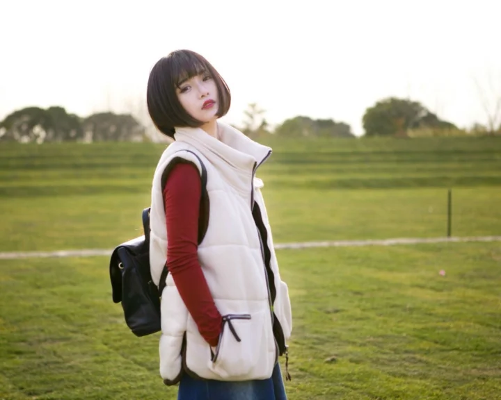 白いベストを着て黒いリュックを背負った女性が、広い芝生の上に立っています。