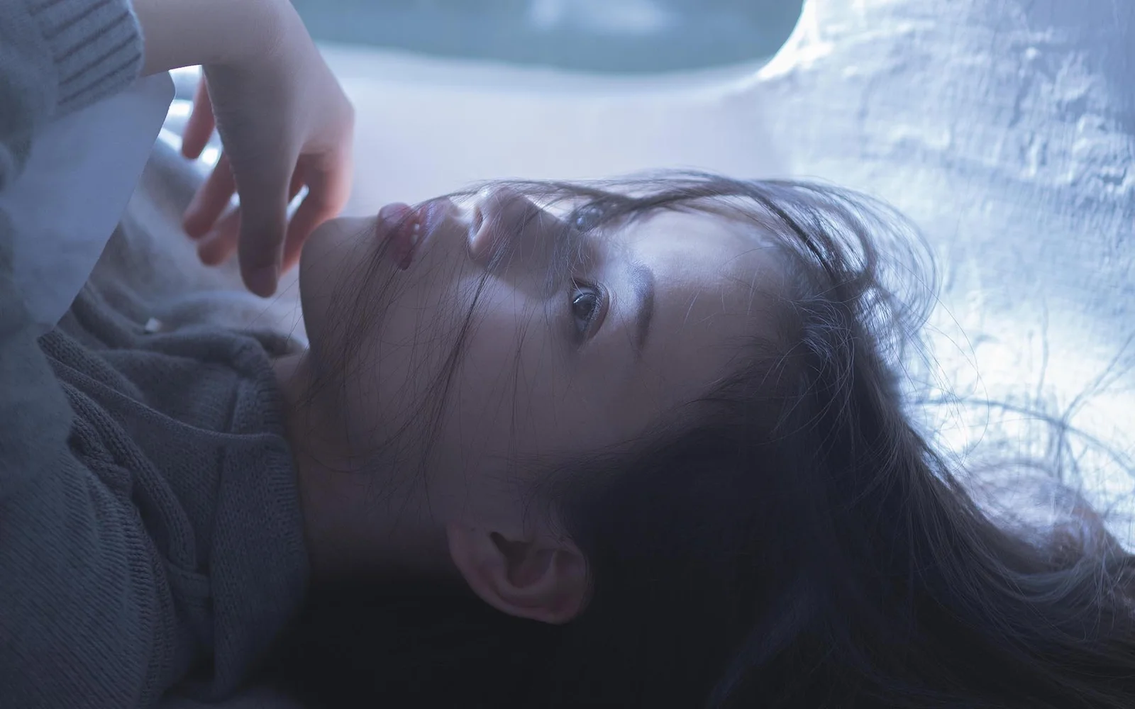 긴 머리의 여성이 차가운 푸른빛 속에 옆으로 누워 얼굴이 화면 대부분을 차지하고 있습니다.