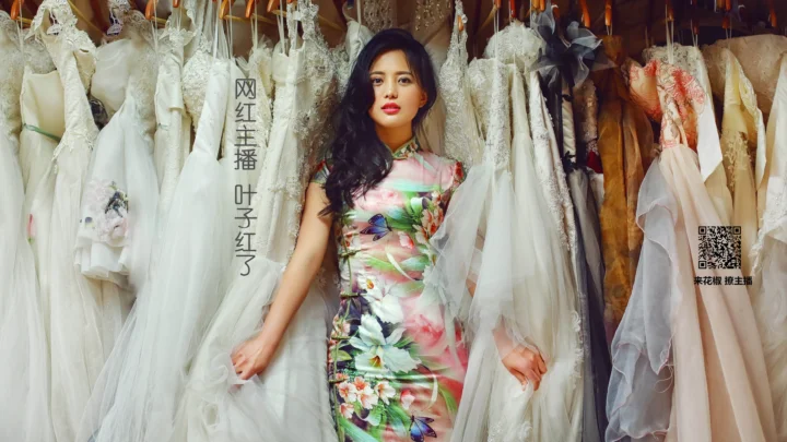 一位成年女性身穿花卉旗袍，站在挂满婚纱和彩色礼服的室内背景前。
