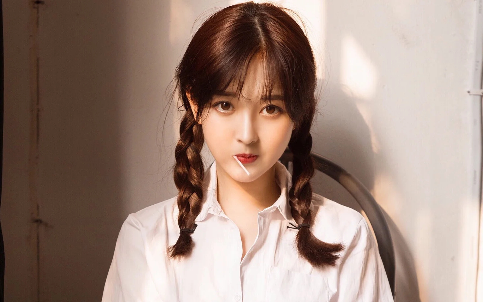 양갈래 땋은 머리에 흰 셔츠를 입은 여성이 따뜻한 실내 조명 속에서 카메라를 바라보고 있다.