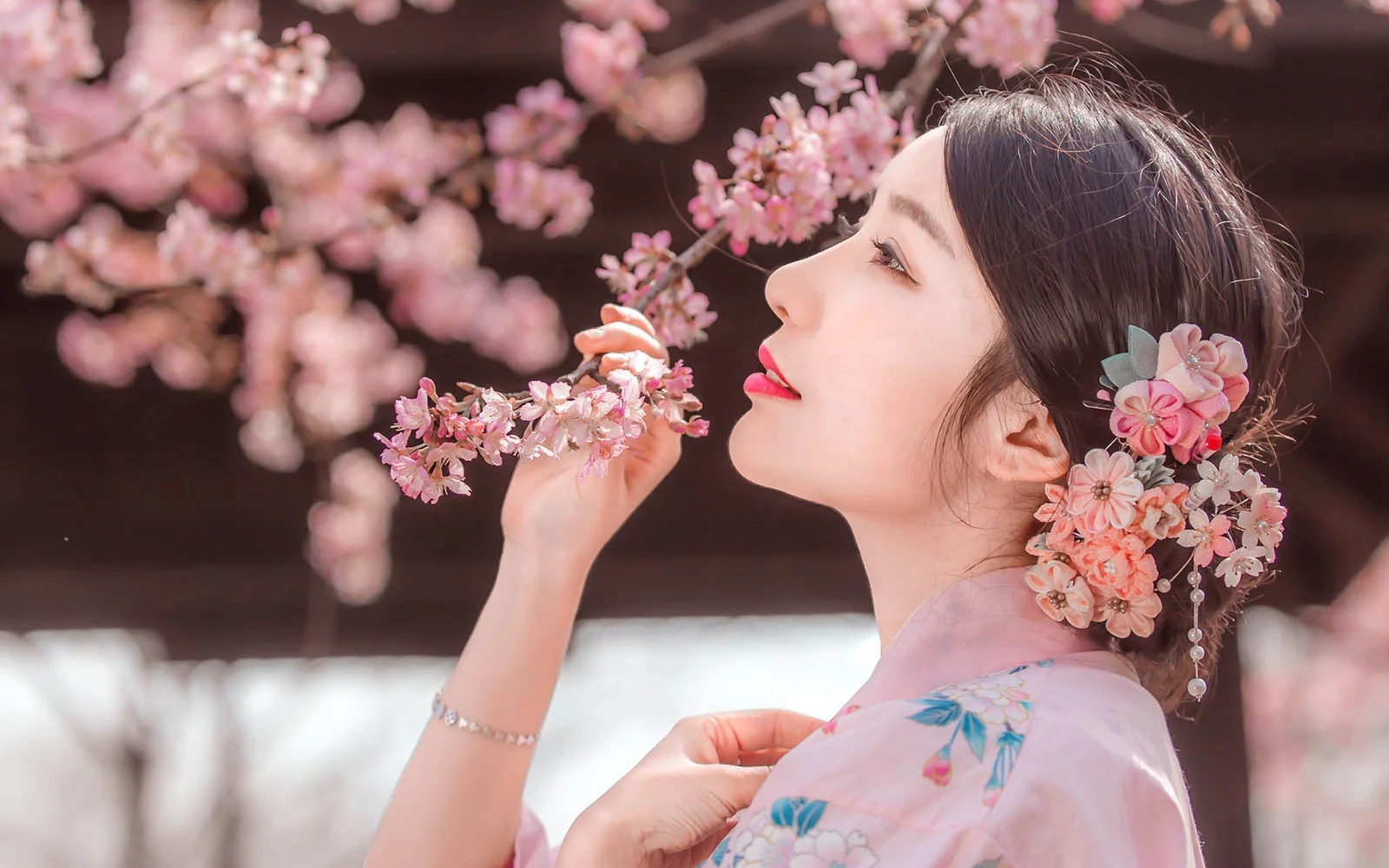 一位成年女性手持樱花枝，发间佩戴花朵饰品，置身粉色樱花之间。
