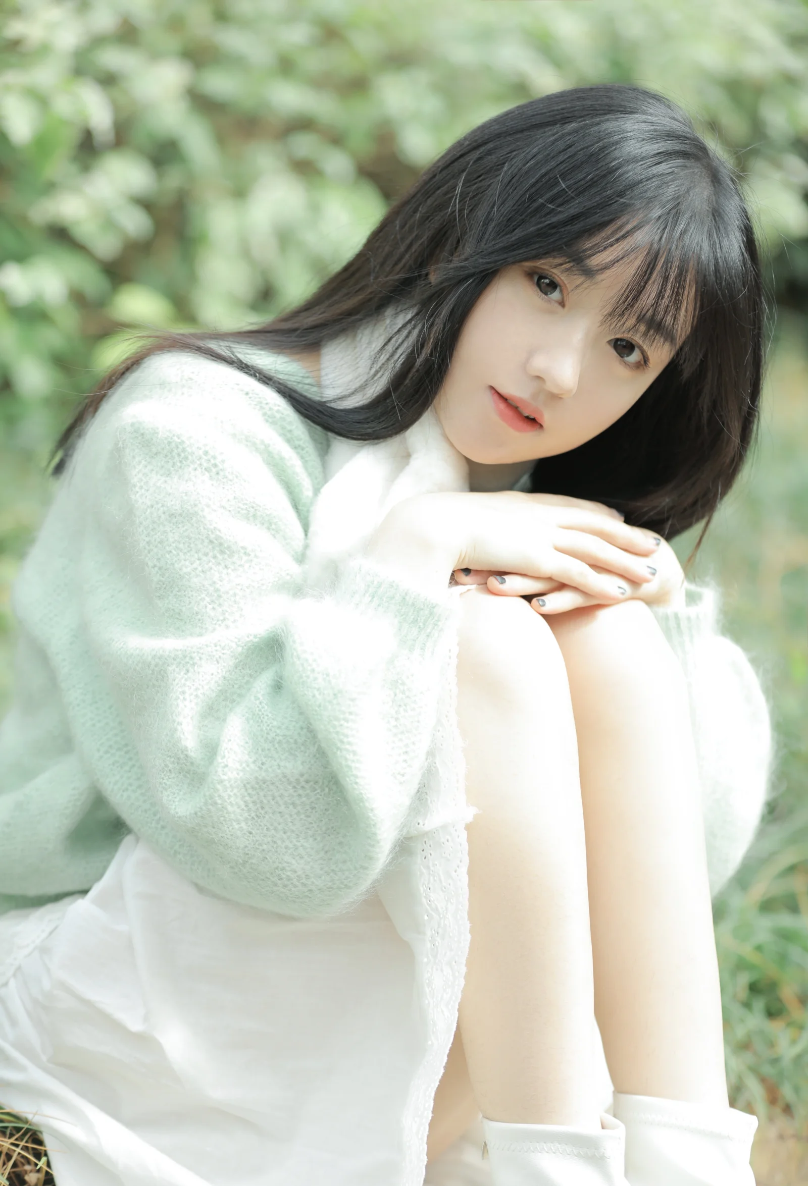 一名黑髮女子在綠色戶外背景前屈膝坐著，雙手交疊放在膝上。