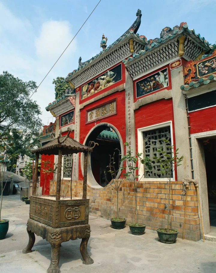 一座红墙装饰华丽的传统建筑矗立在街巷旁，前景摆放着一尊大型香炉。