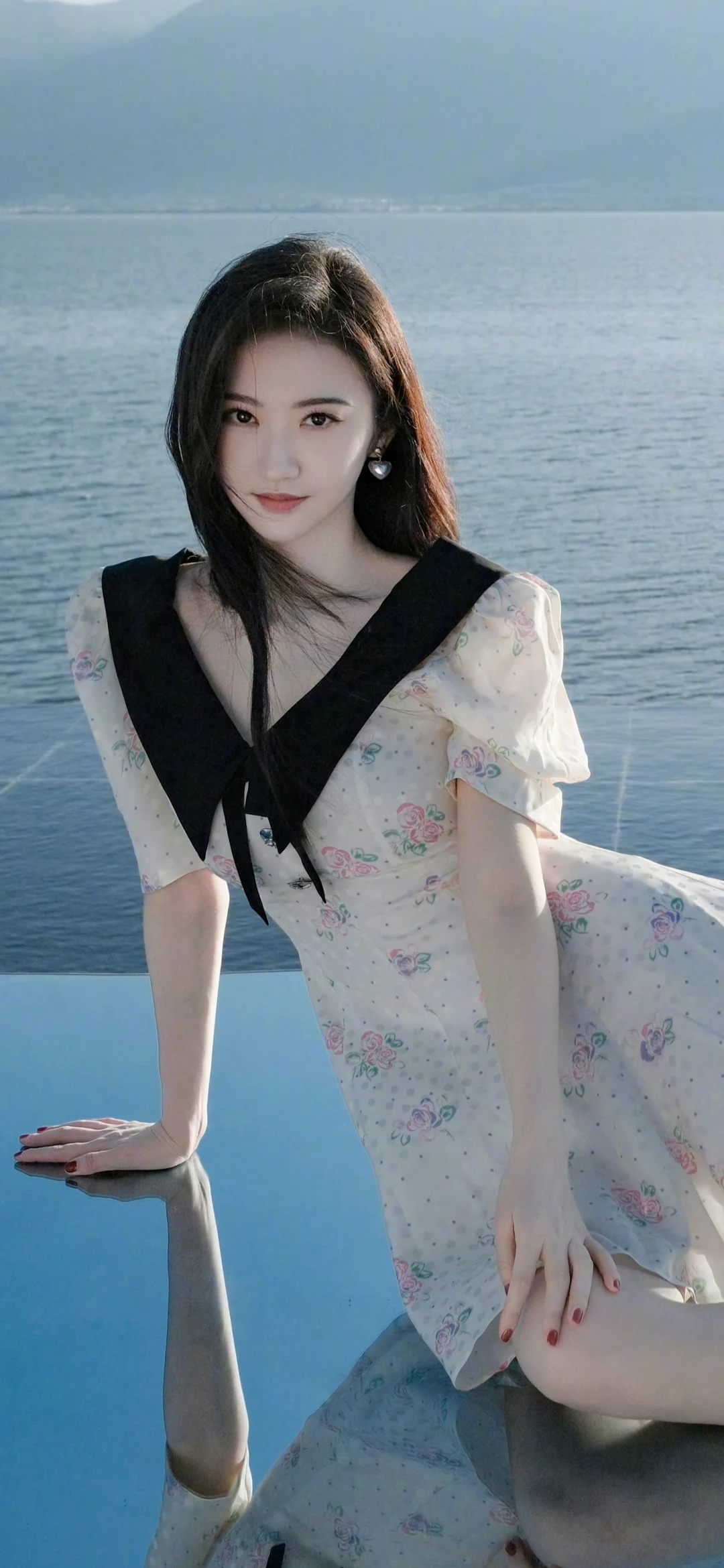一位身穿浅色花卉连衣裙的女性坐在平静湖水旁，远处可见山峦。