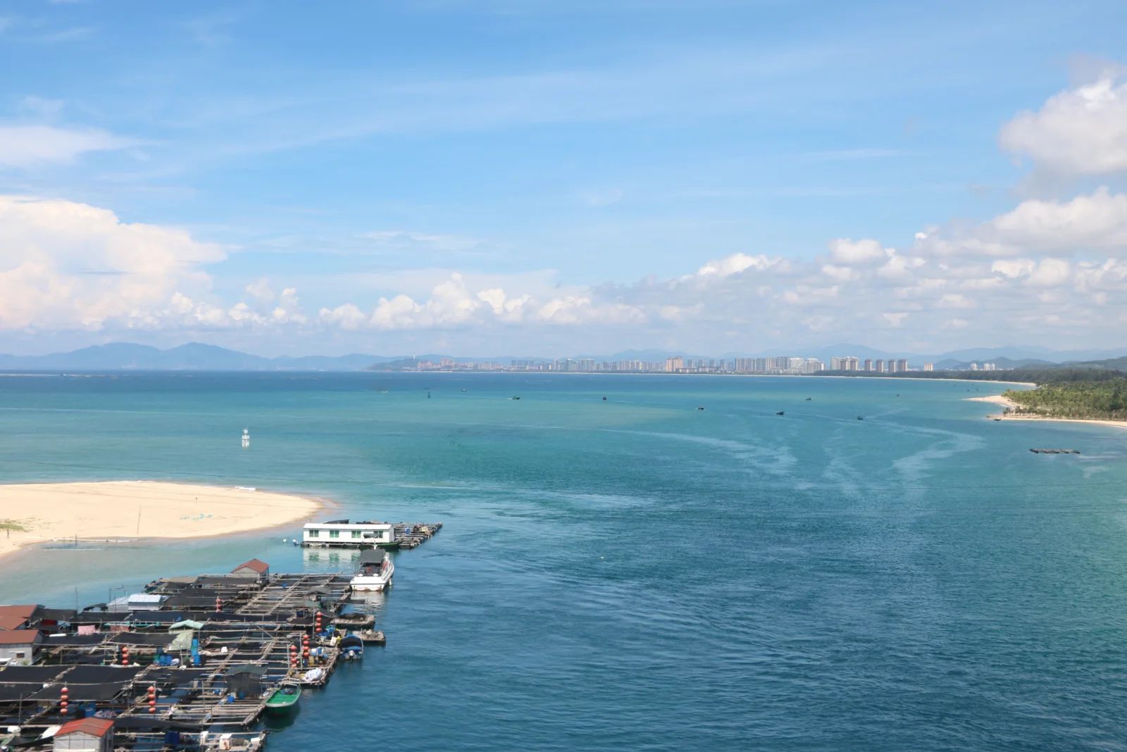画面展现了晴朗天空下的蓝色海湾、沙滩、渔排和远处的城市天际线。
