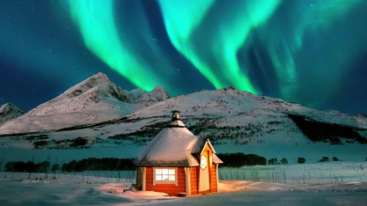 雪地中的木屋坐落在雪山前，夜空中舞动着绿色极光。
