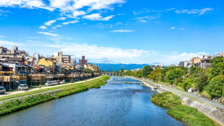一条河流穿过城市，两岸坐落着建筑与绿地，远处可见连绵山脉和蓝天白云。