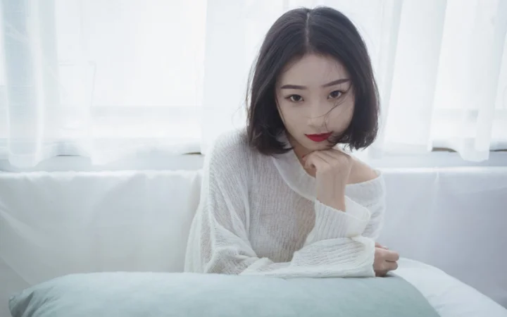 一名成年女性穿着白色针织衫坐在明亮窗边，手托下巴望向镜头。