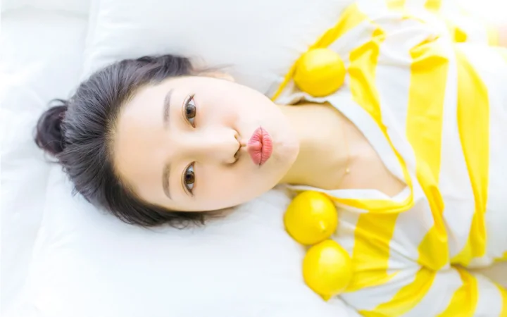一名成年女子躺在白色床铺上，身旁放着几个黄色柠檬。