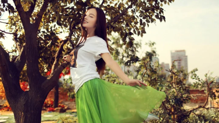 一名身穿白色上衣和绿色长裙的女子站在树下，背景可见树木与远处建筑。