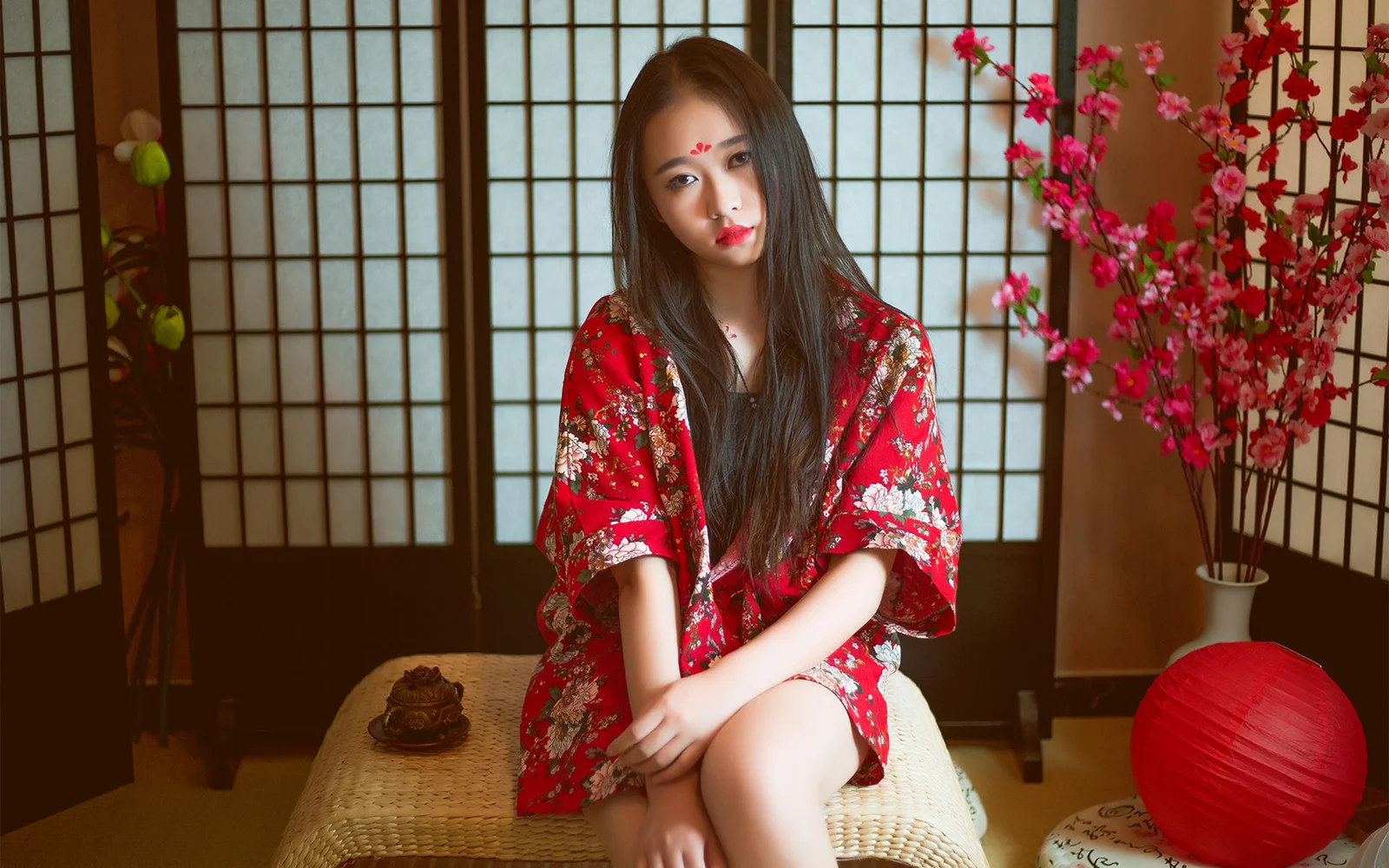 一名成年女性身穿紅色花卉長袍坐在室內藤編椅上，身旁擺放著櫻花枝和紅色燈籠。