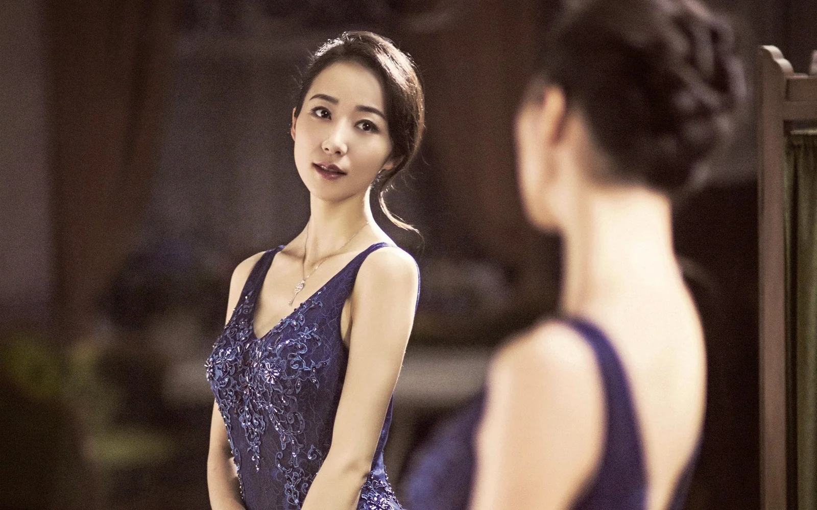 一名成年女子穿着蓝色连衣裙站在镜子前，画面中可见她的倒影。
