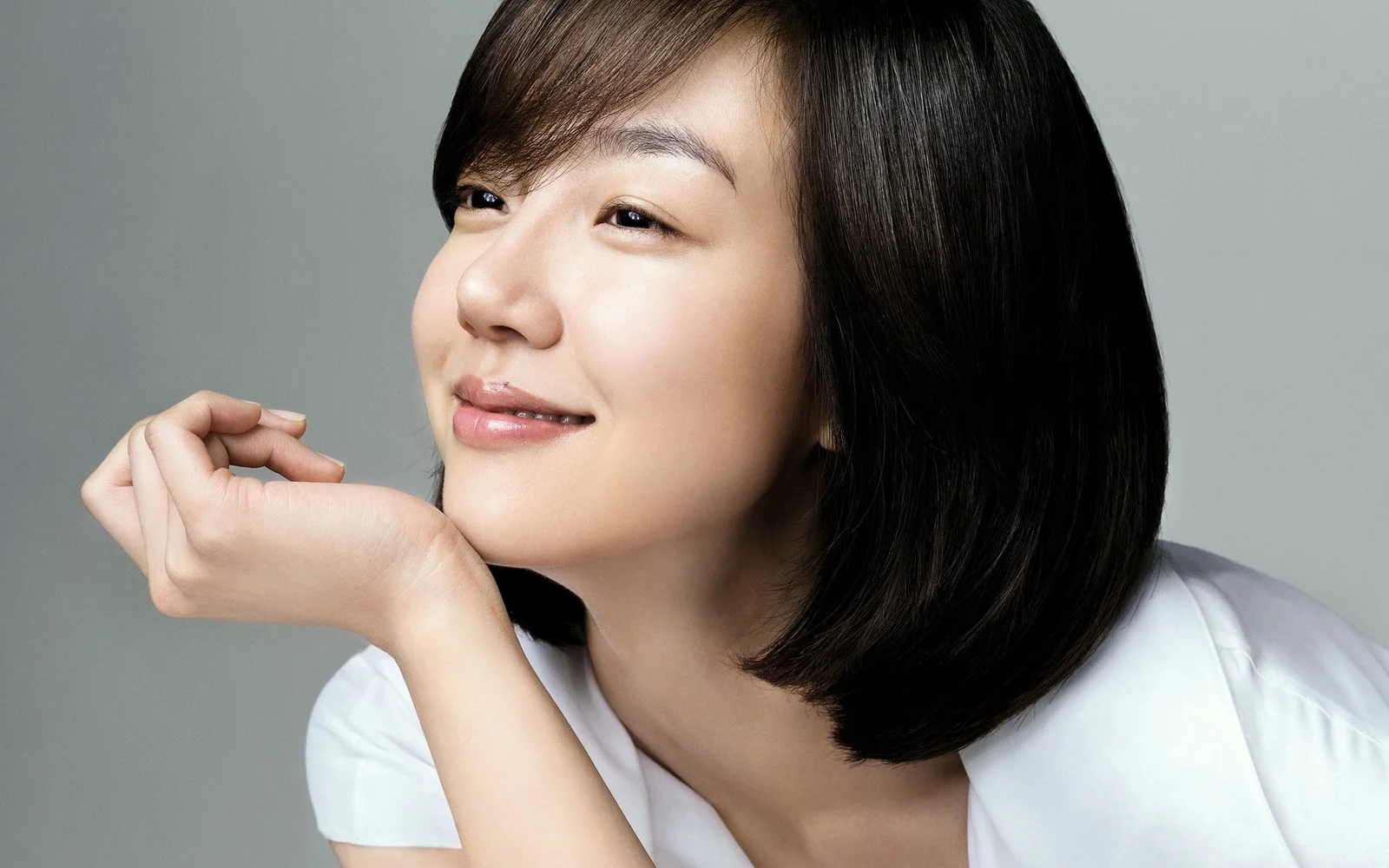 검은 머리의 여성이 흰색 상의를 입고 손으로 턱을 괴며 미소 짓고 있다.