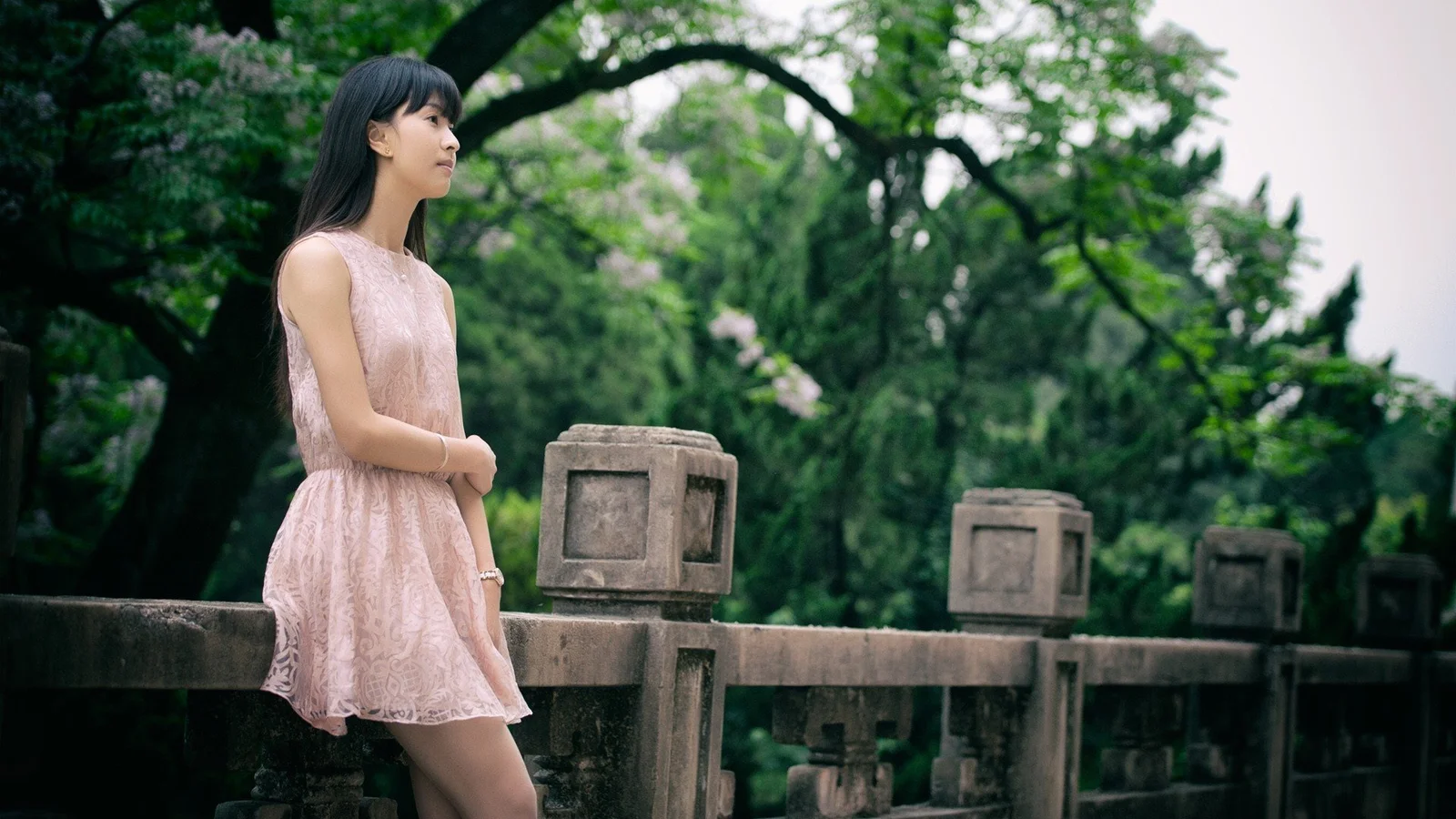 一名穿浅粉色连衣裙的女子站在绿树环绕的石桥旁。