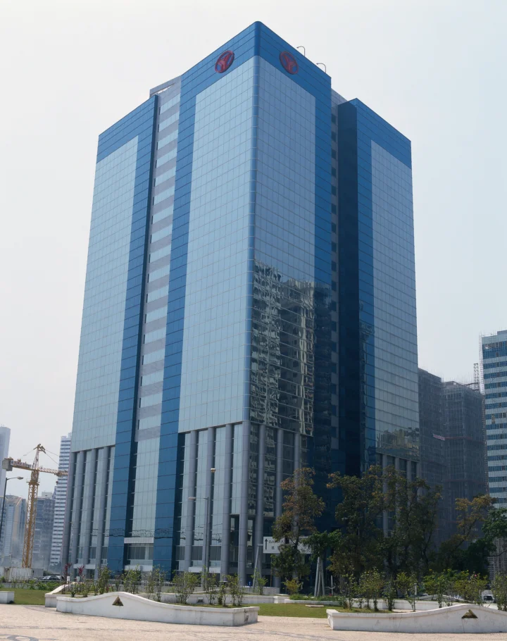 一座高耸的蓝色玻璃办公楼矗立在城市街区中，前方有树木、花坛和施工设施。