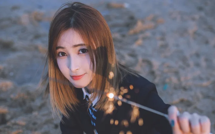 茶色の長い髪の女性が屋外でカメラを見つめ、手前に暖かな玉ボケの光が映っています。