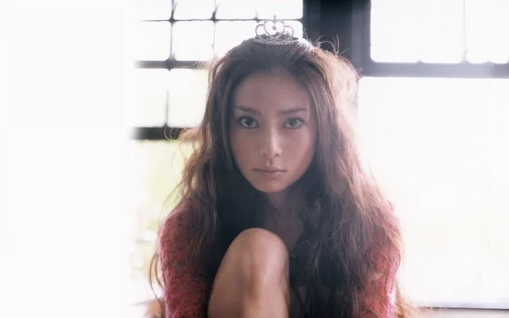작은 티아라를 쓴 긴 머리 여성이 밝은 창가에 앉아 카메라를 바라보고 있습니다.