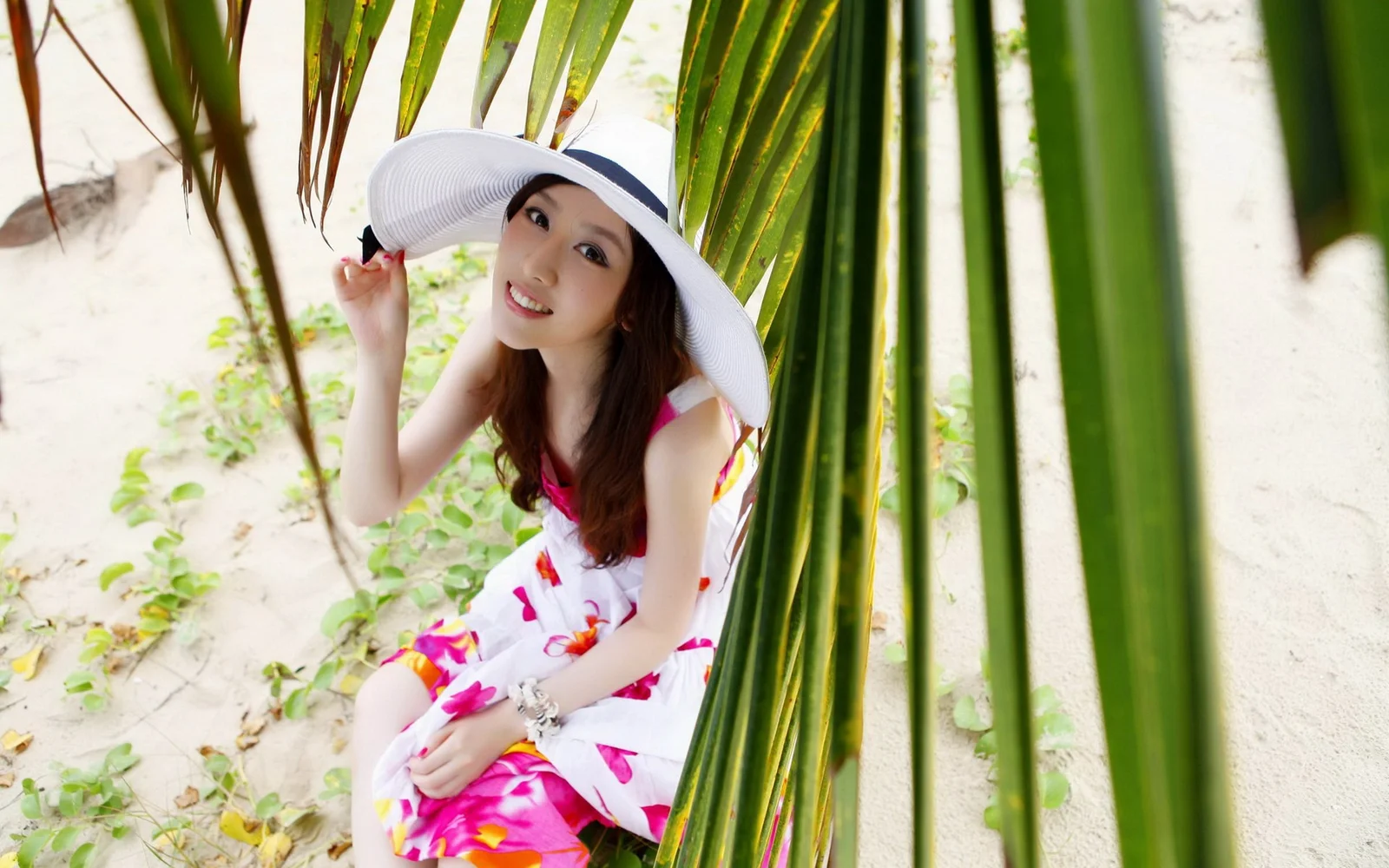 一名戴宽檐帽的女性坐在沙地和热带叶片之间。