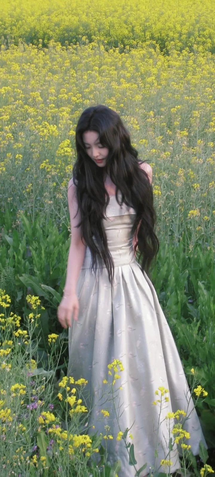 긴 머리의 여성이 연한 색 드레스를 입고 노란 꽃이 핀 들판에 서 있다.