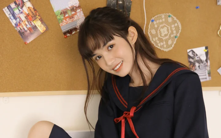一名留着长发的女性身穿深色水手服，在室内软木板前微笑。