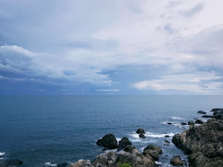 辽阔海面延伸至远方地平线，前景是被海浪拍打的岩石海岸，天空布满层叠云彩。