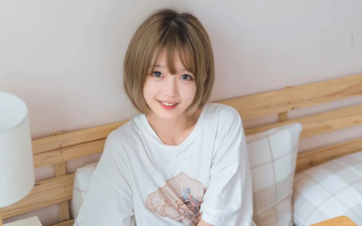 一位短浅棕色头发的女性坐在床上，身穿白色T恤并面带微笑。