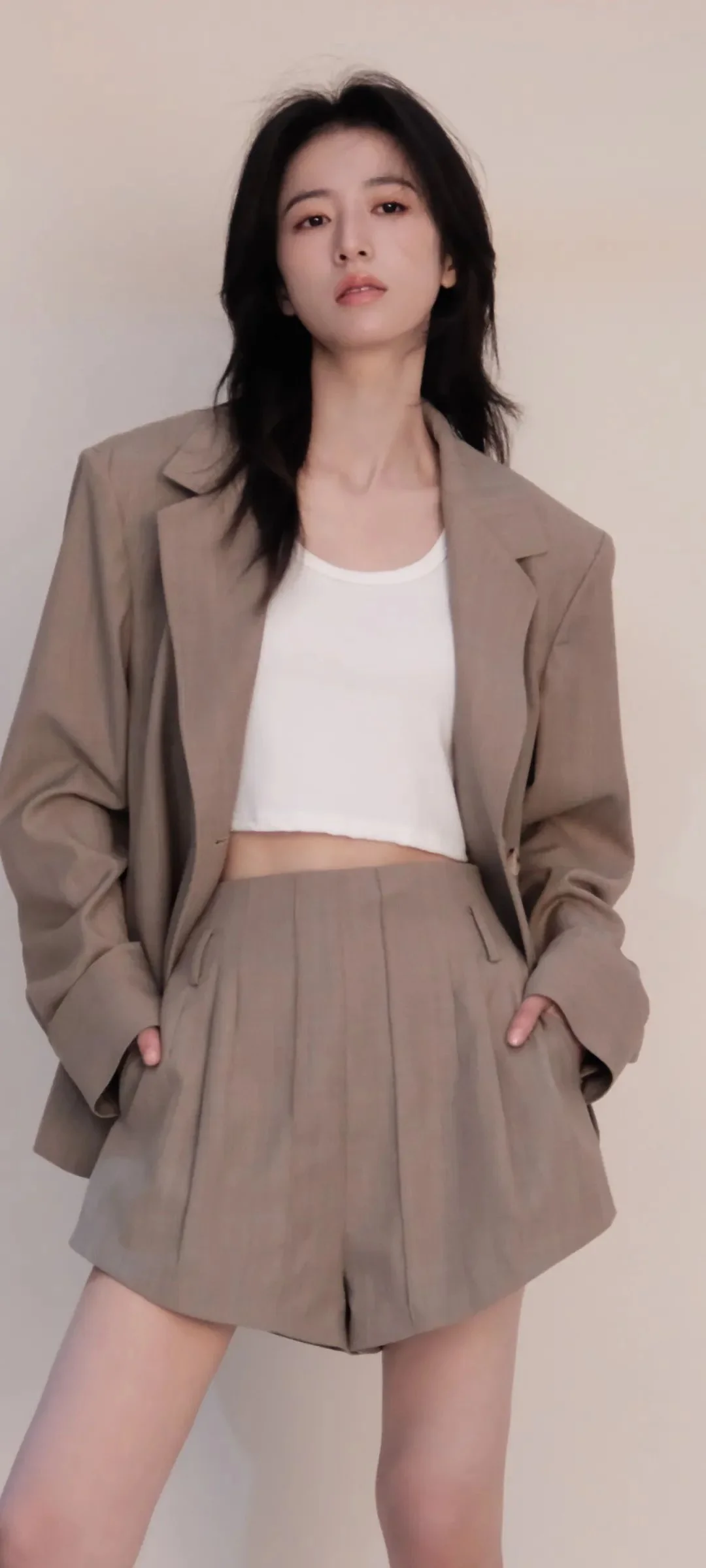 一名成年女性穿着棕色西装外套、白色上衣和短裤站在浅色背景前。