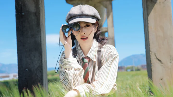 一名戴帽子的成年女性在草地上手持望远镜，身后是蓝天、山景和混凝土柱子。