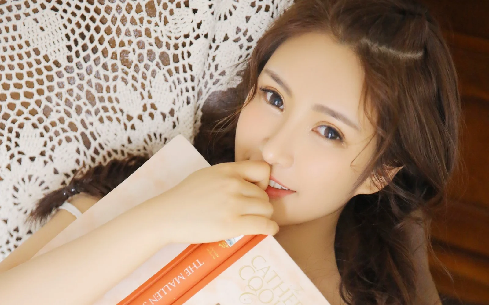 一名棕发女性靠在白色蕾丝背景前，手拿书本并轻触嘴唇。