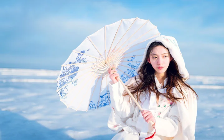 흰 옷을 입은 여성이 바닷가에서 파란색과 흰색 무늬의 양산을 들고 있다.