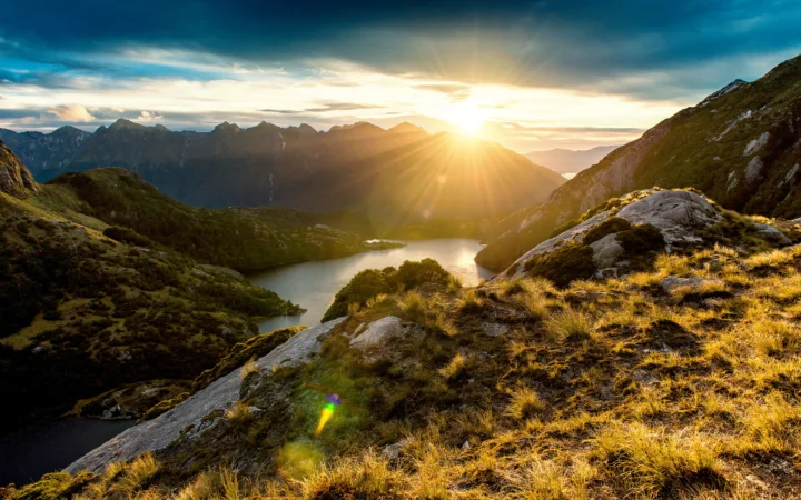夕阳照耀群山与湖泊，前景是金色草地和岩石。