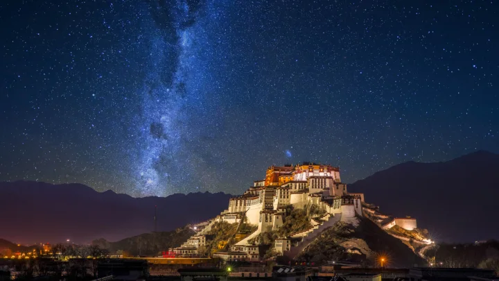 夜空中的银河映照着山上的布达拉宫，远处是连绵的深色山脉。