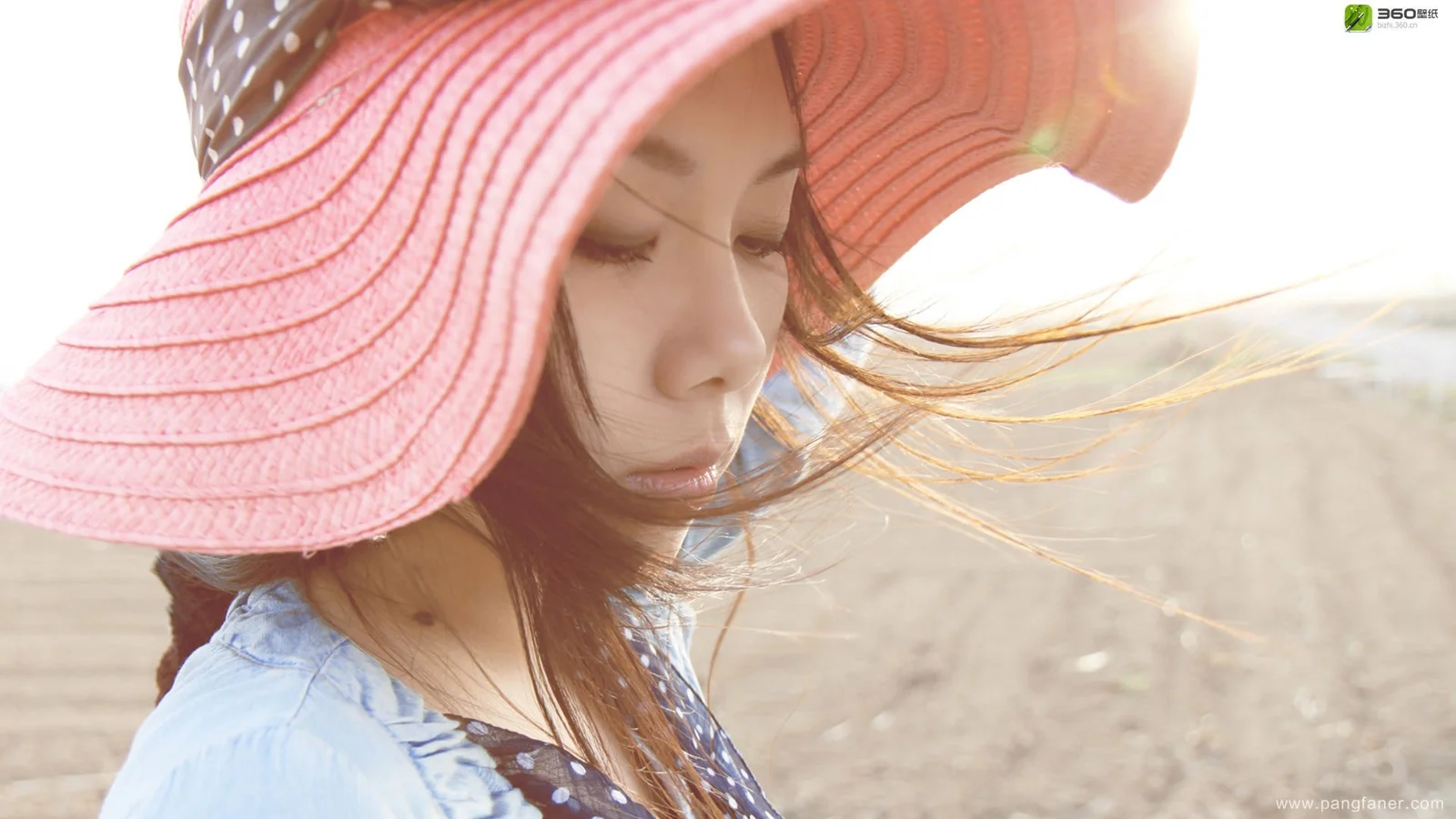 一名戴着粉色遮阳帽的女子在户外低头站立，长发被风吹起。