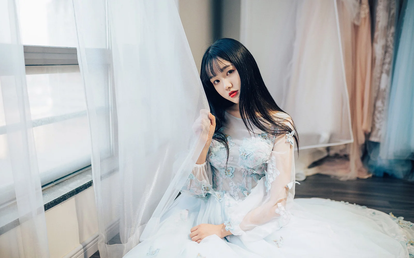 一名身穿白色连衣裙的女子坐在窗边，周围是轻柔的白色窗帘。