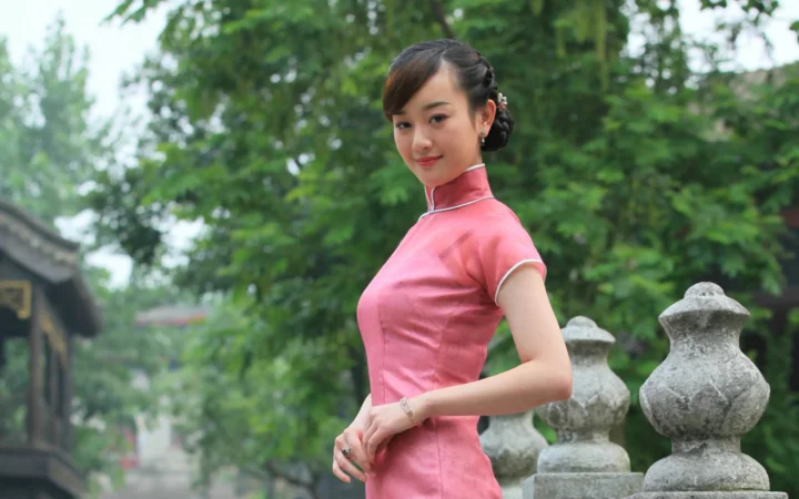 一名穿粉色旗袍的成年女子站在绿树与石雕装饰之间。