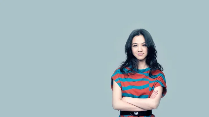 긴 검은 머리의 여성이 줄무늬 셔츠를 입고 팔짱을 낀 채 옅은 청회색 배경 앞에 서 있습니다.