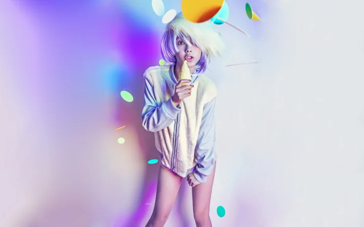 画面中一名短发女性站在紫蓝色与白色的柔和彩光背景前，手持一支浅色物体靠近嘴边。