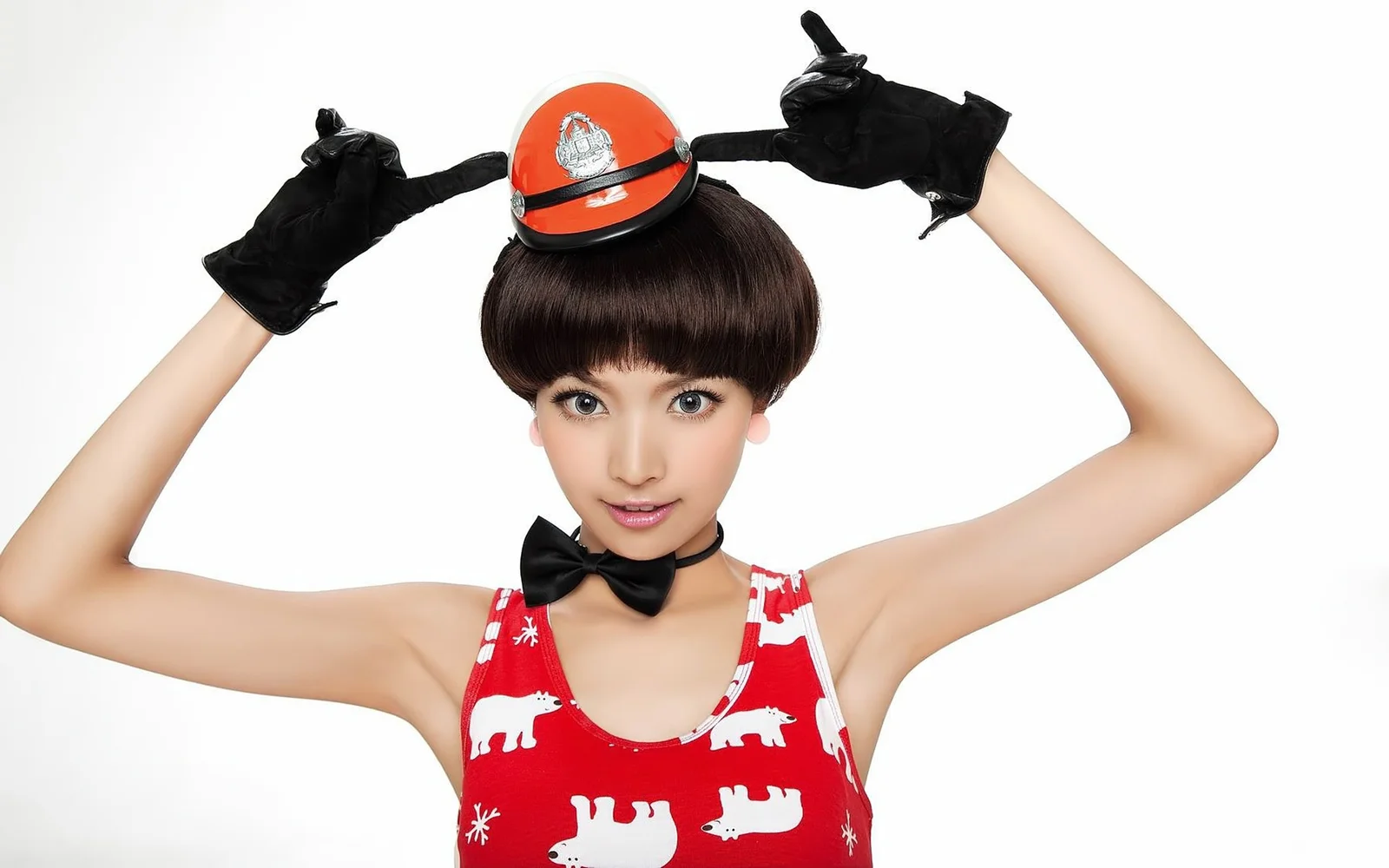 白い背景の前で、赤いヘルメットと黒い蝶ネクタイ、黒い手袋を身につけた女性が両腕を上げている。